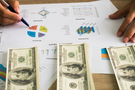 Closeup of Asian male right hand hold ball pen points on business charts under bunch of dollars...