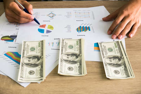 Closeup of Asian male right hand hold ball pen points on business charts under bunch of dollars banknotes for business, finance, tax and people conceptの写真素材