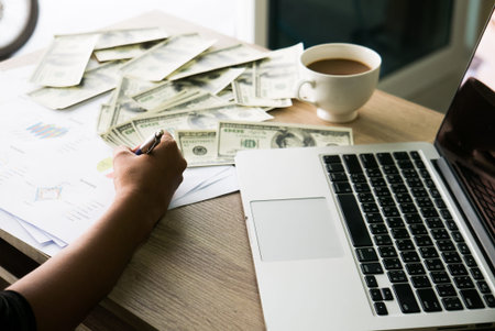 Closeup of Asian female writes something on business charts under bunch of dollars banknotes nearby her labtop and a cop of coffee for business, finance, tax and people conceptの写真素材