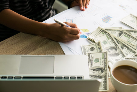 Closeup of Asian female writes something on business charts under bunch of dollars banknotes nearby...