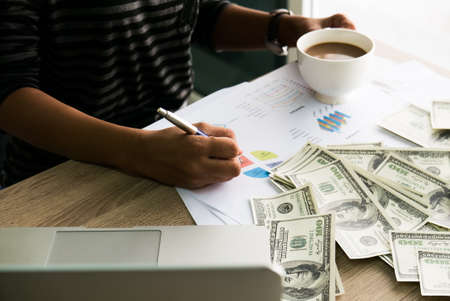Closeup of Asian female writes something on business charts under bunch of dollars banknotes nearby her labtop and a cop of coffee for business, finance, tax and people conceptの写真素材