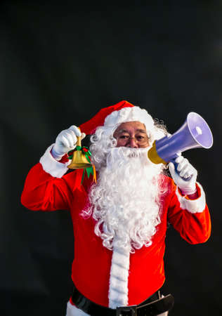 Santa Claus speaks on megaphone on his right hand & ring a bell on his lift hand studio shot on black background for family, giving, season, Christmas, holiday, new year, travel and gathering conceptの写真素材