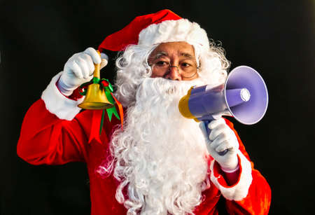 Santa Claus speaks on megaphone on his right hand & ring a bell on his lift hand studio shot on black background for family, giving, season, Christmas, holiday, new year, travel and gathering conceptの写真素材
