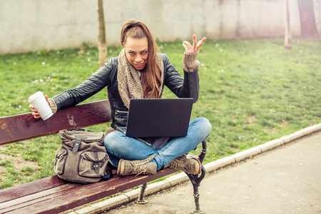 Young female student sitting on park bench, drinking a coffee and working with her laptopの写真素材