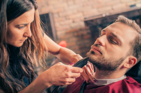 Female barber shaving a client's beard with trimmer in a barber shopの写真素材