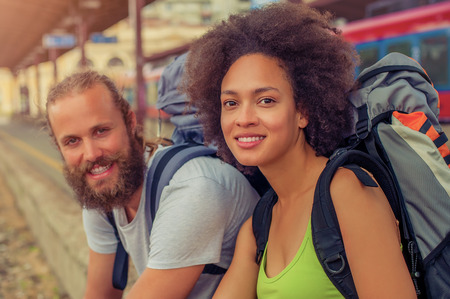 Happy young and beautiful couple of tourists with backpacks sitting at the railway station near the tracks and enjoy their journeyの写真素材