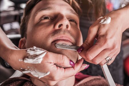 Female barber shaving a client's beard in a barber shop. Close-upの写真素材
