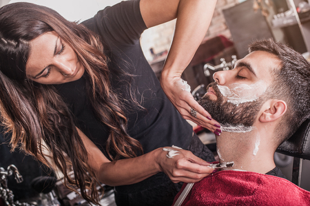 Female barber shaving a client's beard in a barber shop. Close-upの写真素材