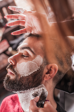 Female barber with shaving brush applied foam to face of client in a barber shop. Close-upの写真素材