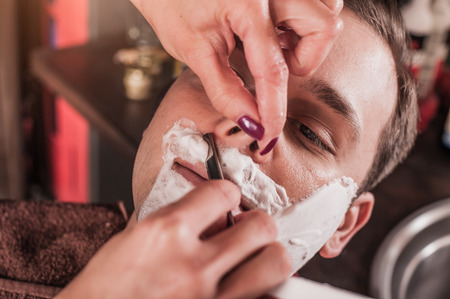 Female barber shaving a client's beard in a barber shop. Close-upの写真素材