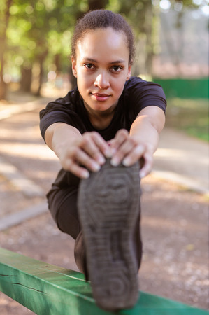 Training outdoors. Fit young woman stretching her legs to warm up. Front angle view.の写真素材