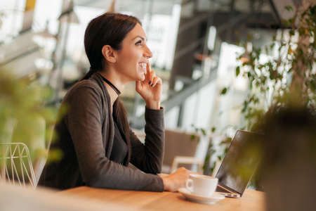 Portrait of smiling young woman drinking a coffee and surfing on internet on her laptop computerの写真素材