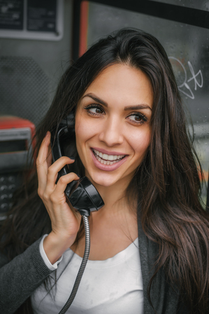 Portrait of happy and smiling woman talking in the retro phone booth on the city streetの写真素材