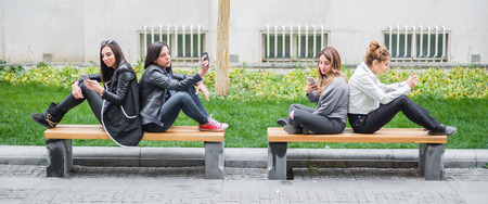 Four girl friends using cellphones on park bench. Internet and social mediaの写真素材