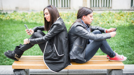 Two girl friends using cellphones on park bench. Internet and social media