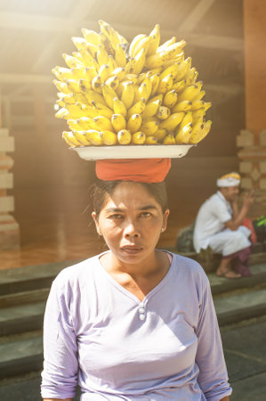 BALI, INDONESIA - 13. APRIL 2018. Balinese woman carrying bananas on her head outside a temple. Day editorial shotのeditorial素材