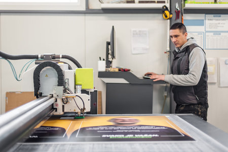 Printshop worker technician works on large CNC computer numerical control printing and cutting...