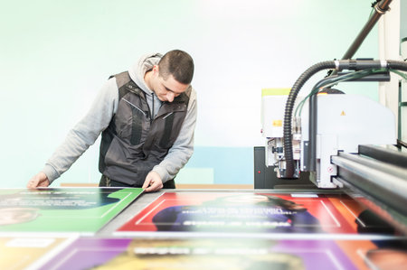 Printshop worker technician works on large CNC computer numerical control printing and cutting...