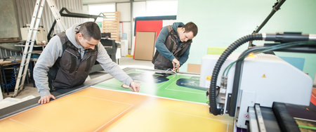 Two printshop worker technicians works on large CNC computer numerical control printing and cutting...