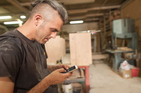 Carpenter artisan resting in workshop and scrolling on smartphone. Handsome handicraftsman texting message and tapping on phoneの写真素材