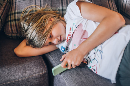 Teen girl checking social media holding smartphone, using mobile phone app playing game, relaxing on sofa at home. Emotional isolation and depressionの写真素材