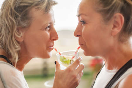 Two cheerful girlfriends drink beverage from one glass using straws, enjoying cold beverage on a summer day outdoorsの写真素材
