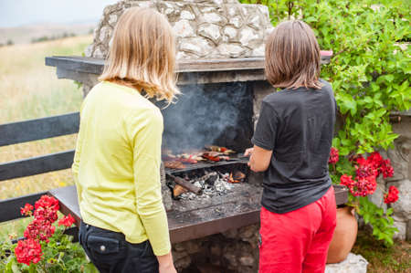Children grilling meat. Boy and girl making barbecue on the grill on nature. Family camping and enjoying BBQの写真素材