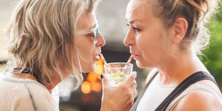 Two cheerful girlfriends drink beverage from one glass using straws, enjoying cold beverage on a summer day outdoorsの写真素材