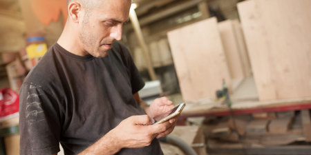 Carpenter artisan resting in workshop and scrolling on smartphone. Handsome handicraftsman texting...