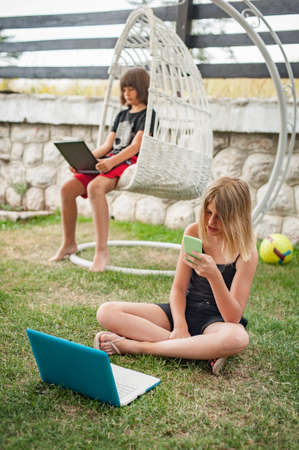 Social alienation of teenagers. Two children using laptop and smartphone while resting in backyard. Young people addiction to technology trendsの写真素材