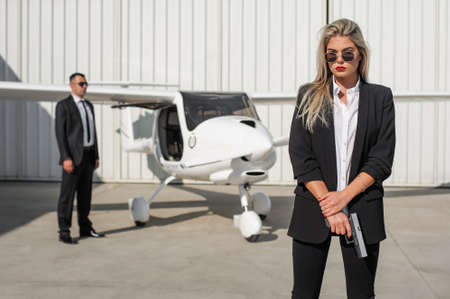 Beautiful professional female spy agent bodyguard with gun securing airplane on private airport. Security police woman in civilian black suit. Close security protectionの写真素材