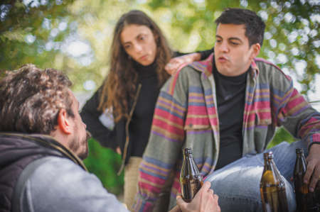 Group of female and male young friends drinks beer from bottle on wooden bench with table in forest park outdoor. Woman and man casual lifestyle conceptの写真素材