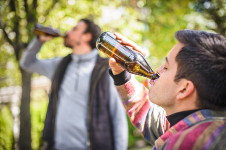 Two men chugging beer from bottle on wooden bench with table in forest park outdoor. Express...