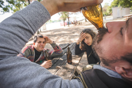 Young group of friends, female and male, chugging beer from bottle on wooden bench with table in...