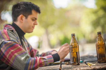 Lonely man drinks beer from bottle alone and thinks about the meaning of life. Wooden bench with table in forest park outdoor. Real life sceneの写真素材