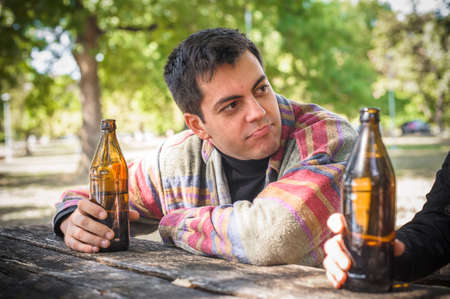 Moody man enjoys life and drinks beer from bottle. Wooden bench with table in forest park outdoor. Real life sceneの写真素材