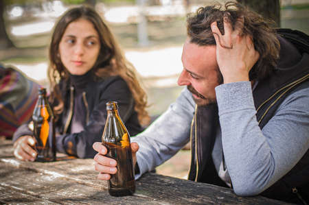 Smiling cheerful young couple, boyfriend and girlfriend, drinks beer from bottle on wooden bench with table in forest park outdoor. Real life scene. Woman and man casual lifestyle conceptの写真素材