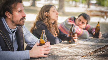 Group of three unhappy young friends, female and male, sit together and drinks beer from bottle on wooden bench with table in forest park outdoor. Real life scene. Social depression among young peopleの写真素材