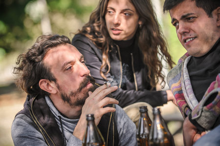 Group of relaxed and smiling happy young friends, female and male, smoking cannabis marijuana ganja or hashish joint in public park outdoor. Young people smoke thc grass. Real life scene conceptの写真素材