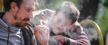 Two young friends smoking cannabis marijuana ganja or hashish joint in public park outdoor. Two men...
