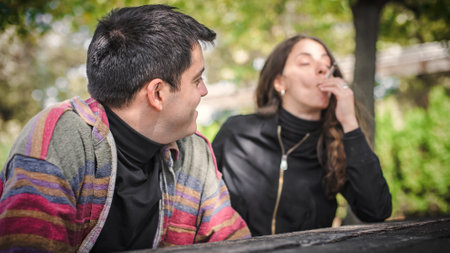 Young relaxed and smiling happy young couple, woman and man, smoking cannabis marijuana ganja or hashish joint in public park outdoor. Female and male smoke thc green grass together. Real lifeの写真素材