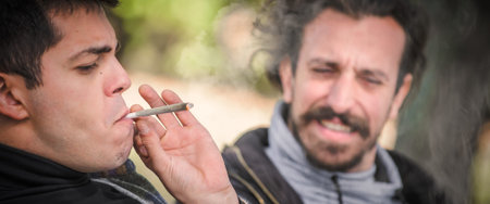 Two young friends smoking cannabis marijuana ganja or hashish joint in public park outdoor. Two men...
