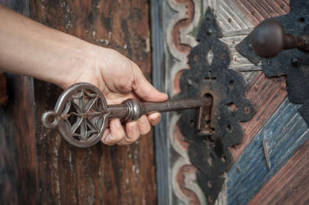 Closeup detail view of man holds a huge massive medieval church key in his hand and unlocks or locks a large wooden door. Secret and mysteryの写真素材