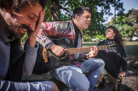 Positive and relaxed young friends play guitar and singing together while sitting on park bench and enjoy in beautiful romantic summer dayの写真素材