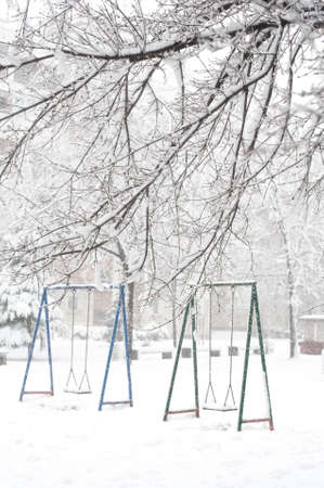 Frozen white children playground and swings for swinging in the snowy city winter landscape. Winter snow outdoors view.の写真素材