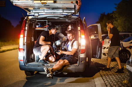 Behind scene improvisation. Cameraman film crew team from trunk of van car shooting night film scene on the street outdoor. Group filmmaking set productionの写真素材