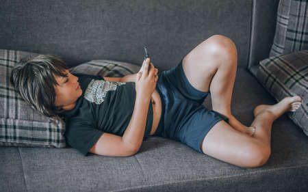Teen boy checking social media holding smartphone, using mobile phone app playing game, relaxing on sofa at home. Emotional isolation and depressionの写真素材