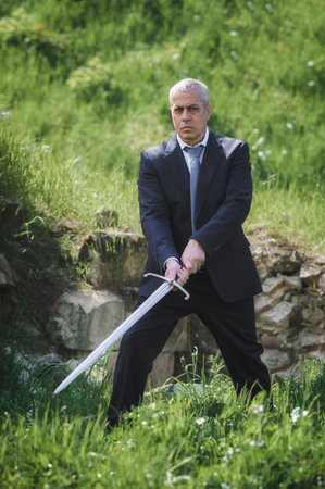 Portrait of handsome businessman model man in a modern casual suit with a large medieval sword....