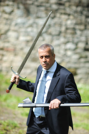 Portrait of handsome businessman model man in a modern casual suit with a large medieval sword....