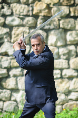 Portrait of handsome businessman model man in a modern casual suit with a large medieval sword....
